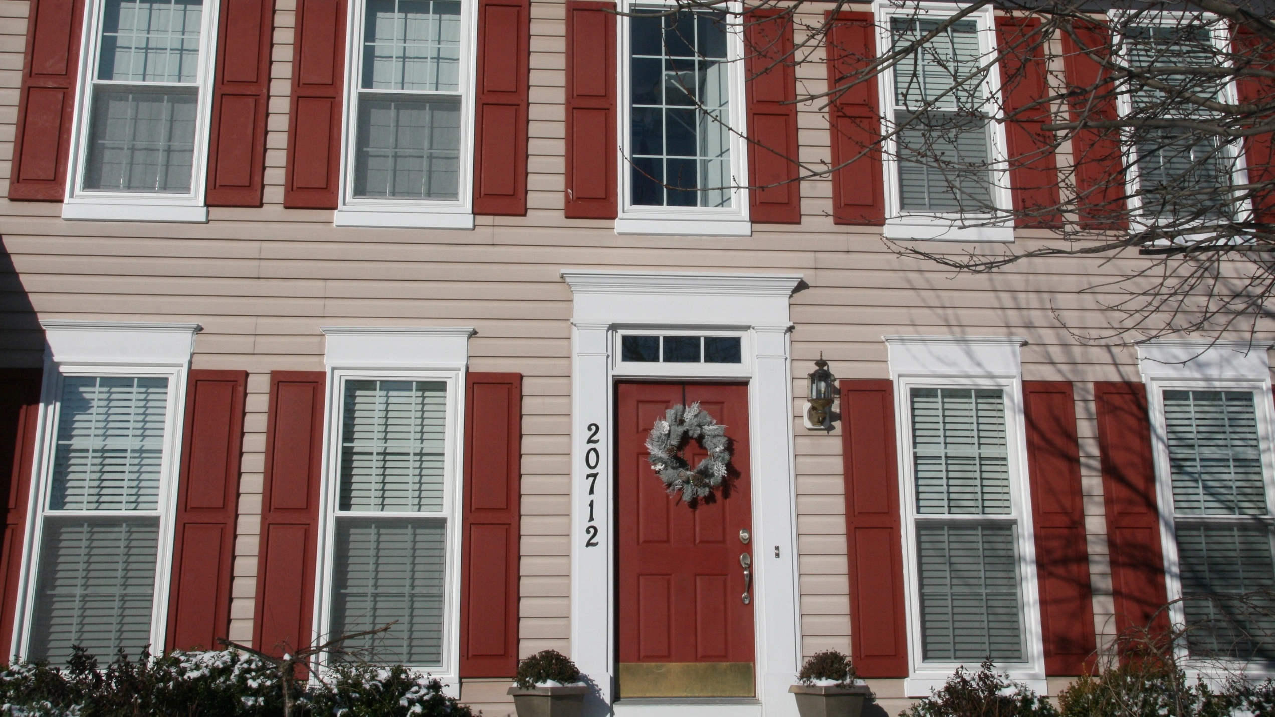A two-story home with beige vinyl siding, red shutters and entry door, white windows, and blinds in the windows